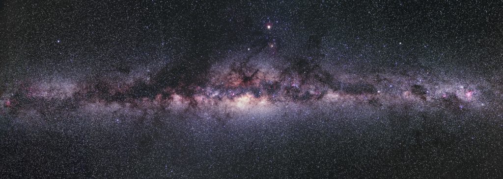 Otthonunk, a Tejút galaxis panorámája. Középen a magvidék, sárgás, öregebb csillagok fényével, a szélek felé vörösesen derengő hidrogén ködökkel, sötét porfelhőkkel és kékes, fiatal csillagokkal, csillaghalmazokkal.